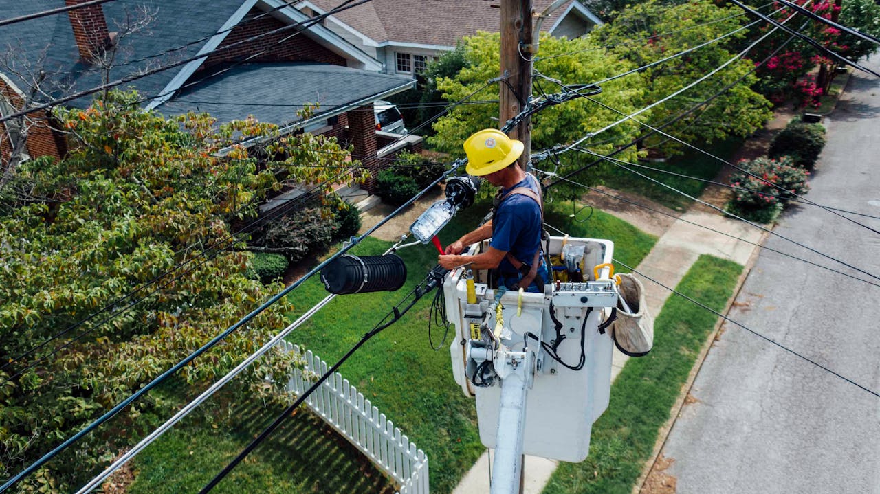 hero-img-01 Electrician in a bucket lift repairing power lines from a utility pole in a suburban neighborhood.